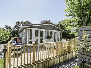 A wooden deck with chairs and a table outside a building surrounded by trees at Orchard Lodge in Llanengan near Abersoch