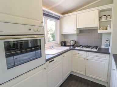 A kitchen with white cabinets a built in oven a gas stove a sink a toaster a kettle and shelves with containers at Orchard Lodge in Llanengan near Abersoch