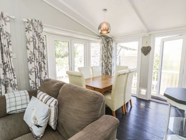 A dining area with a wooden table and white chairs next to a brown sofa and floral curtains at Orchard Lodge in Llanengan near Abersoch
