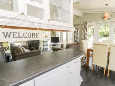 A kitchen counter with a welcome sign overlooking a living room with sofas and a television and a dining area with a wooden table and cream chairs at Orchard Lodge in Llanengan near Abersoch