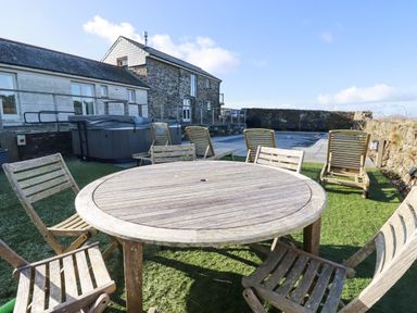 An outdoor patio area with a round wooden table and chairs on grass near a stone building and a swimming pool at Penpethy Farm in Tintagel