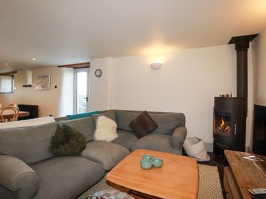 A living room with a gray sectional sofa a wood burning stove a wooden coffee table and dining chairs at Penpethy Farm in Tintagel