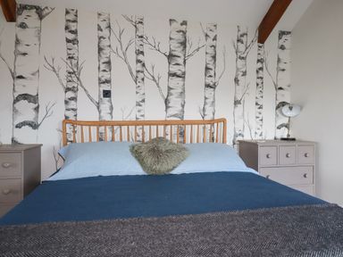 A bedroom with a wooden bed frame blue bedding two gray bedside tables and wallpaper with birch tree designs at Penpethy Farm in Tintagel