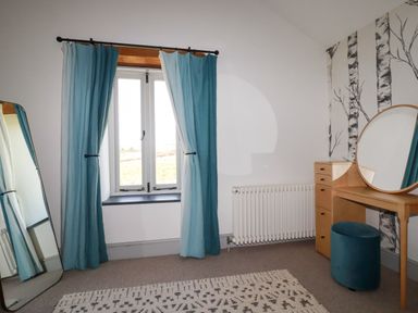 A bedroom with a window with blue curtains a full length mirror a patterned rug and a wooden vanity with a round mirror and blue stool at Penpethy Farm in Tintagel