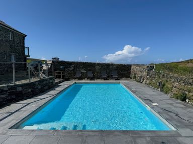 An outdoor swimming pool with lounge chairs along a stone wall and a house with patio furniture nearby at Penpethy Farm in Tintagel