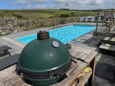 An outdoor swimming pool with blue water and lounge chairs beside it and a green ceramic grill on a wooden table at Penpethy Farm in Tintagel