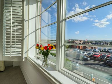 A window with white shutters and a vase of red and yellow flowers on the sill overlooking a parking lot and buildings at Above & Beyond in Whitby