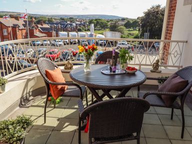 A balcony with a round table and four chairs with cushions overlooking a town with red brick buildings and parked cars at Above & Beyond in Whitby