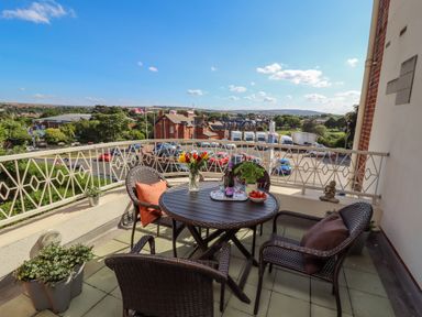 A balcony with a round table and four chairs with cushions overlooking parked cars and buildings at Above & Beyond in Whitby
