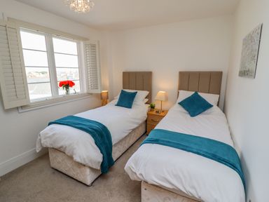 A bedroom with two single beds with blue throws and pillows a wooden nightstand with two lamps and a window with shutters and flowers at Above & Beyond in Whitby