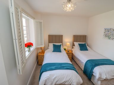 A bedroom with two single beds side by side with teal throws and pillows a bedside table with a lamp and flowers on the window sill at Above & Beyond in Whitby