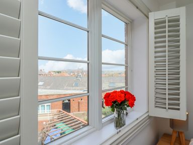 A window with white shutters partly open and a vase with red roses on the windowsill at Above & Beyond in Whitby