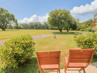 Two wooden chairs on a patio facing a grassy area with trees and a pathway at Lodge 16 in Lanteglos near Camelford
