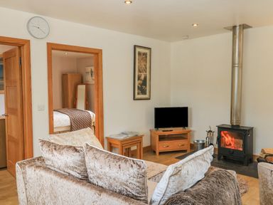 A living room with a grey sofa a wood-burning stove a television on a wooden stand and a doorway leading to a bedroom at The Mews in Bovey Tracey near Chudleigh Knighton