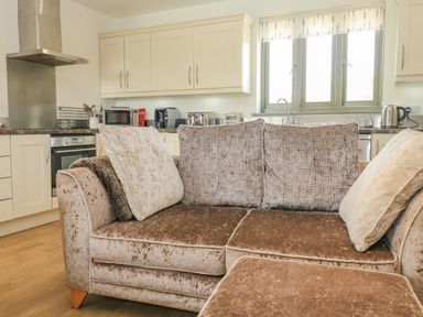 A kitchen area with beige cabinets and appliances behind a brown velvet sofa with cushions at The Mews in Bovey Tracey near Chudleigh Knighton