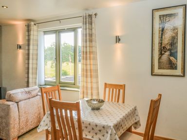 A dining area with a wooden table covered by a patterned cloth and four wooden chairs near a window with curtains and a framed picture on the wall at The Mews in Bovey Tracey near Chudleigh Knighton