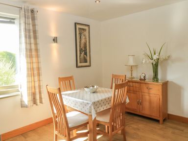A dining area with a wooden table covered with a white patterned tablecloth and four wooden chairs a wooden sideboard with a lamp clock and flowers a window with checkered curtains and a framed picture on the wall at The Mews in Bovey Tracey near Chudleigh Knighton