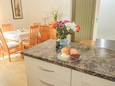 A kitchen counter with flowers and scones and a dining table with chairs and a cupboard in the background at The Mews in Bovey Tracey near Chudleigh Knighton