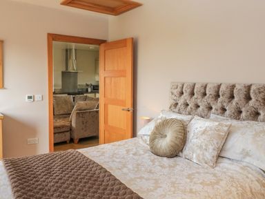 A bedroom with a bed featuring decorative pillows and a blanket with an open door showing a kitchen and sofa at The Mews Bovey Tracey near Chudleigh Knighton