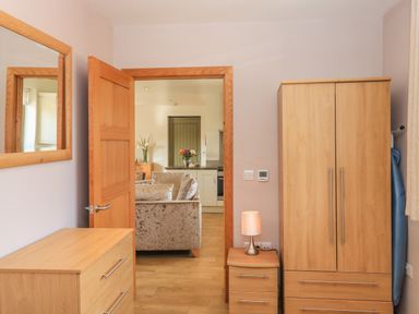 A bedroom with wooden dresser, wardrobe, bedside table with lamp and an open door showing a living area at The Mews in Bovey Tracey near Chudleigh Knighton