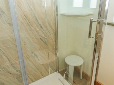 A shower area with marble-like wall panels and a glass door with a metal handle and a white stool inside at The Mews in Bovey Tracey near Chudleigh Knighton