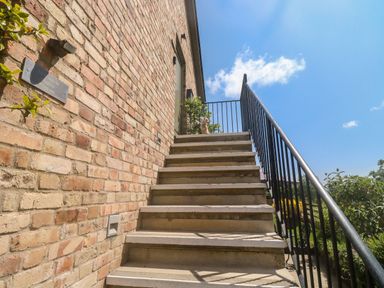 An outdoor staircase with black metal railing leading to a door on a brick building with plants and a small dog statue at The Mews in Bovey Tracey near Chudleigh Knighton