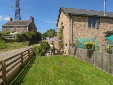A garden with outdoor table and chairs under an umbrella next to a brick house with greenhouses and a wooden fence at The Mews in Bovey Tracey near Chudleigh Knighton