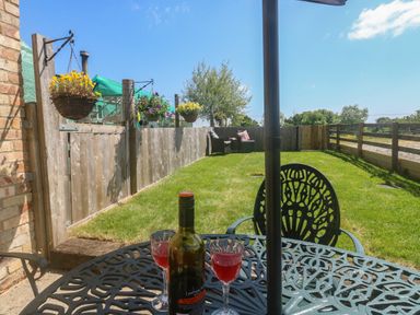 A garden with a metal table and two glasses of red drink on it a bottle of wine hanging flower baskets a wooden fence and chairs at The Mews in Bovey Tracey near Chudleigh Knighton