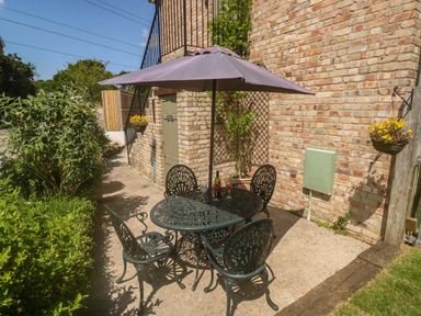 An outdoor patio area with a metal table and four chairs under a large umbrella next to a brick wall with plants and flowers at The Mews in Bovey Tracey near Chudleigh Knighton