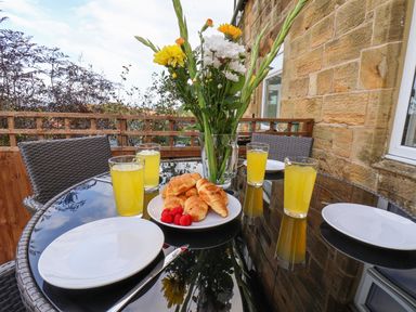 An outdoor glass table with four plates four glasses of yellow drink a plate of croissants and raspberries and a vase of flowers at Maypole Cottage in Fylingthorpe