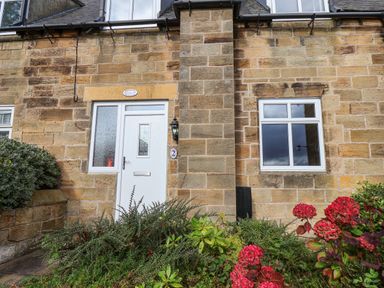 The front exterior of a stone cottage with a white door and window surrounded by plants and red flowers at Maypole Cottage in Fylingthorpe