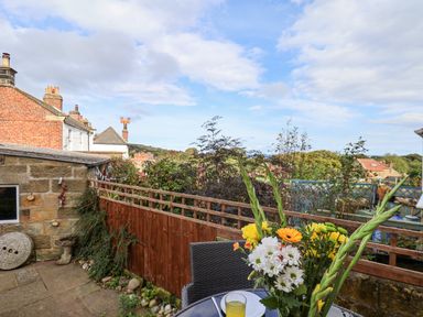A patio with a table and flowers in a garden with houses and trees in the background at Maypole Cottage in Fylingthorpe