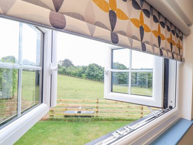 A view through an open double window with patterned blinds showing a grassy field and trees beyond a wooden fence at Sunflower Sparnon Gate near Illogan