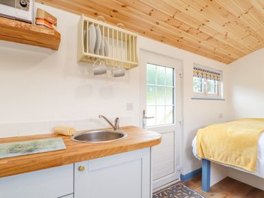 A kitchen area with a sink wooden countertop plates and mugs on a rack a small window a bed with a yellow blanket and a door at Sunflower Sparnon Gate near Illogan