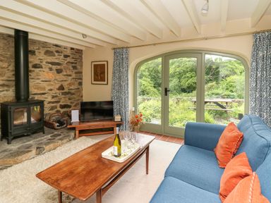 A living room with a wood stove next to a stone wall and large glass doors showing greenery outside at Taibach in Dinas Cross