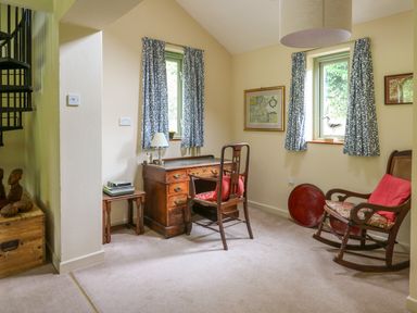 A room with a wooden desk and chair a rocking chair with a cushion and two windows with patterned curtains at Taibach in Dinas Cross