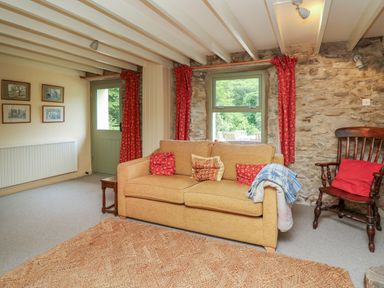 A living room with a beige sofa red cushions wooden chair and stone wall at Taibach in Dinas Cross