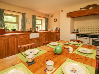 A kitchen with wooden cabinets a dining table set with plates cups and a green teapot at Taibach in Dinas Cross