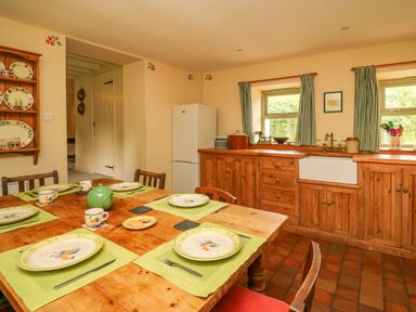 A kitchen with a wooden dining table set with plates and cups and wooden cabinets with a white refrigerator and windows with green check curtains at Taibach in Dinas Cross