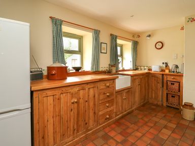A kitchen with wooden cabinets tiled floor and two windows with curtains at Taibach in Dinas Cross