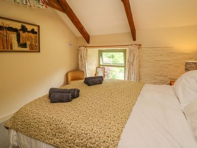 A bedroom with a bed covered by a patterned quilt and rolled towels on top a window with floral curtains and exposed wooden beams at Taibach in Dinas Cross