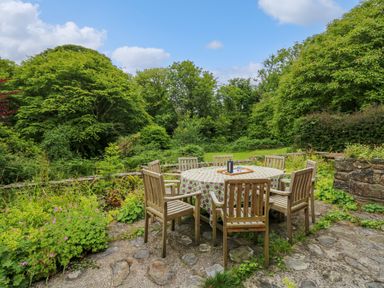 An outdoor patio with a round table covered by a tablecloth and wooden chairs surrounded by greenery at Taibach in Dinas Cross
