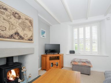 A living room with a wood burning stove a TV on a wooden cabinet a gray ottoman a wooden coffee table and a window with white shutters at Quay House in Charlestown