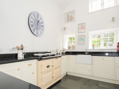 A kitchen with white cabinets black countertops a beige oven a farmhouse sink two windows and a large wall clock at Quay House in Charlestown