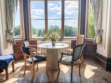 A round table with a flower vase and four chairs near large windows with curtains at Nethercott House in Iddesleigh near Hatherleigh