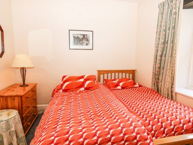 A bedroom with two single beds covered with red patterned bedspreads a wooden nightstand with a lamp a window with floral curtains and a framed picture on the wall at Nethercott House in Iddesleigh near Hatherleigh
