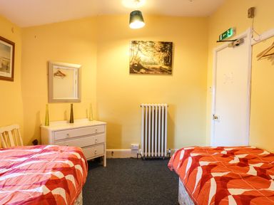 A room with two beds with red patterned covers a white chest of drawers with vases a radiator and a door at Nethercott House in Iddesleigh near Hatherleigh