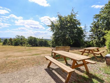 Two wooden picnic tables and a bench on gravel next to a grass field with trees in the background at Nethercott House in Iddesleigh near Hatherleigh