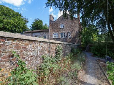 A path beside a stone wall with plants and a stone building in the background at Nethercott House in Iddesleigh near Hatherleigh