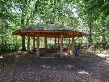 An outdoor wooden shelter with a grass roof surrounded by trees at Nethercott House in Iddesleigh near Hatherleigh